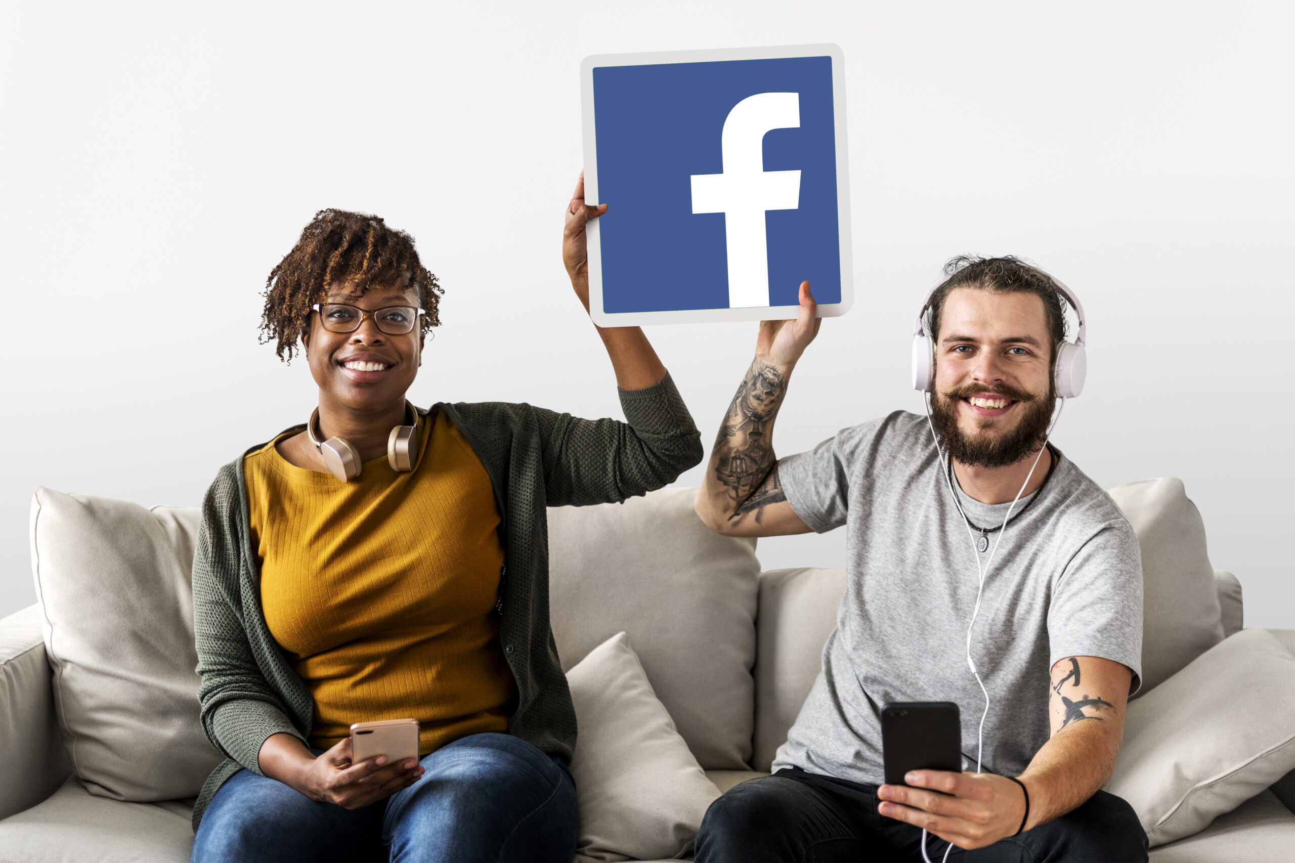 Two smiling people sitting on a couch holding a large Facebook logo and smartphones.