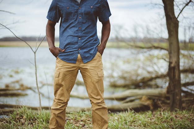 A man stands outdoors near a serene lake, wearing a blue button-up shirt and mustard-colored cargo pants.