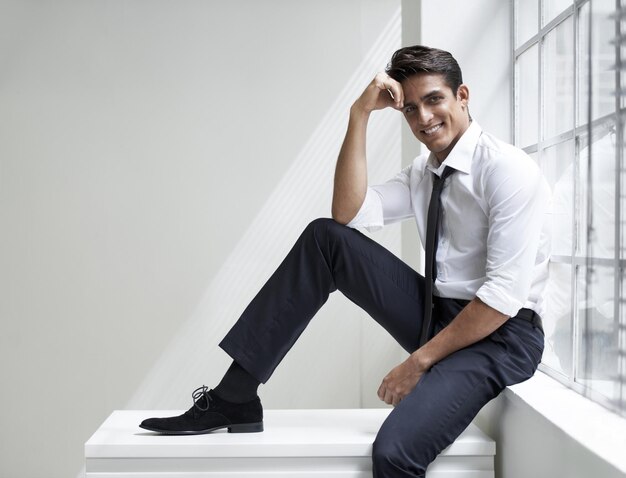 Smiling man in white shirt and tie sitting by a window in modern formal office wear.