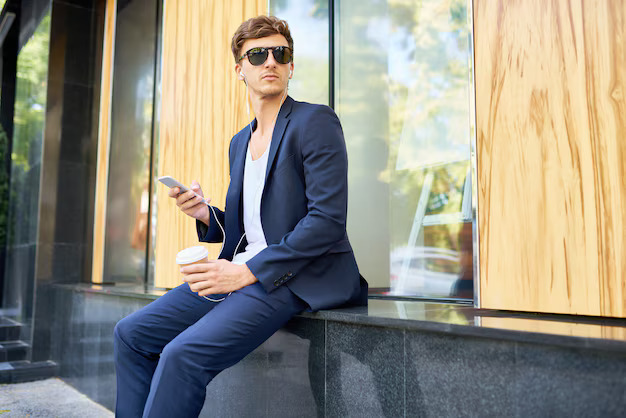 Stylish man in navy suit and sunglasses holding coffee and phone outside an office building.

